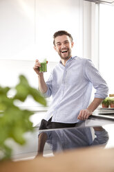 Porträt eines lachenden jungen Mannes mit grünem Smoothie in seiner Küche - MFRF000540