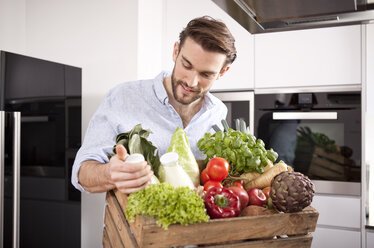 Porträt eines jungen Mannes mit einer Holzkiste mit frischem Gemüse und zwei Milchflaschen in seiner Küche - MFRF000546