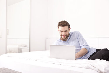 Smiling young man lying on a hotel bed using laptop - MFRF000558