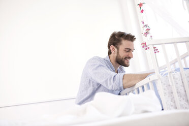 Happy young man sitting beside baby cot looking down - MFRF000560