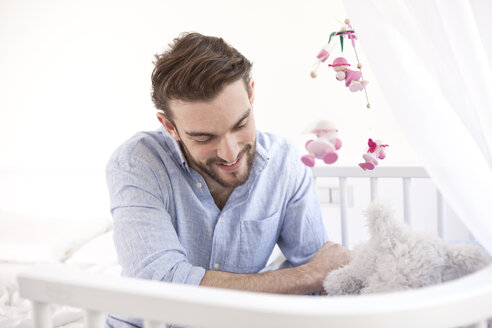 Glücklicher junger Mann mit Plüschtier, der neben einem Babybettchen sitzt und nach unten schaut - MFRF000561