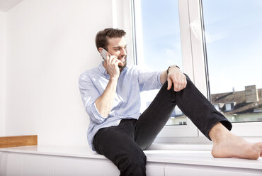 Junger Mann sitzt auf einem Sideboard und telefoniert mit seinem Smartphone, während er durch das Fenster schaut - MFRF000566