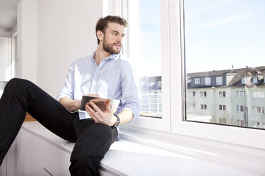 Young man with mini tablet sitting on sideboard at home looking through window - MFRF000569