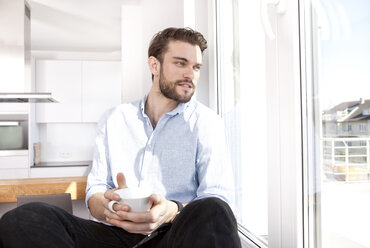 Junger Mann mit Kaffeetasse auf dem Fensterbrett sitzend und durch das Fenster schauend - MFRF000572