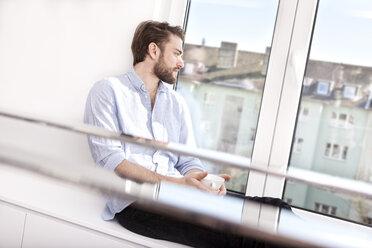 Young man with coffee cup sitting on sideboard looking through window - MFRF000574