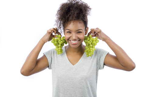 Porträt einer lächelnden jungen Frau, die grüne Weintrauben wie Ohrringe vor einem weißen Hintergrund hält, lizenzfreies Stockfoto