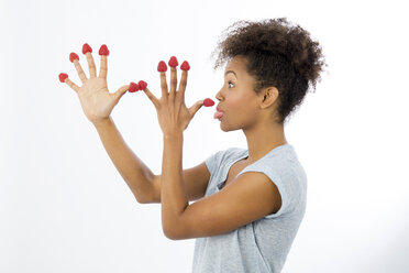 Young woman with raspberries on her fingertips pulling funny faces in front of white background - GDF000978
