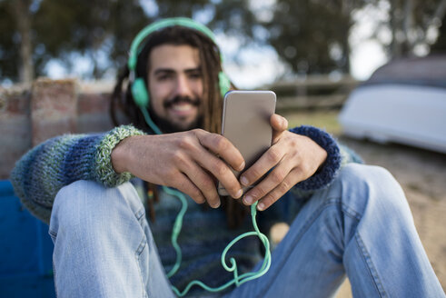 Junger Mann mit Dreadlocks hört Musik und benutzt das Smartphone - KIJF000296