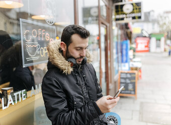 UK, London, man with headphones standing in front of window display looking at his smartphone - MGOF001684