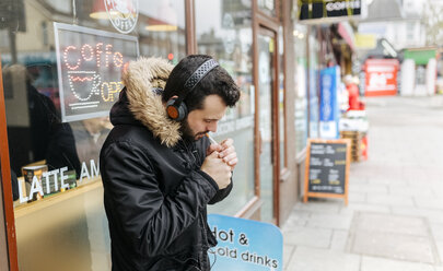 UK, London, man with headphones standing in front of window display lighting a cigarette - MGOF001685