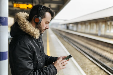 Mann mit Kopfhörern steht auf dem Bahnsteig und benutzt sein Smartphone - MGOF001686