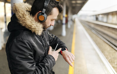 Mann mit Kopfhörern steht auf einem Bahnsteig und schaut auf seine Smartwatch - MGOF001687