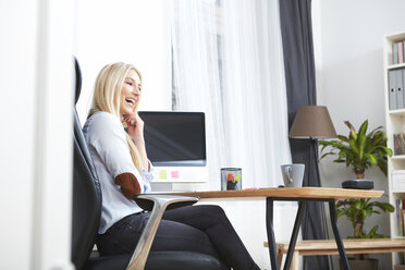 Lachende blonde Frau, die in ihrem Büro zu Hause am Schreibtisch sitzt - SEGF000487