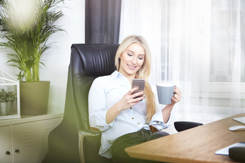 Porträt einer lächelnden blonden Frau, die mit einer Tasse Kaffee an ihrem Schreibtisch sitzt und auf ihr Smartphone schaut, lizenzfreies Stockfoto