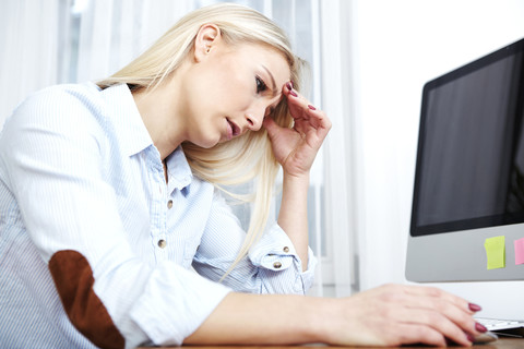 Überforderte blonde Frau sitzt am Schreibtisch, lizenzfreies Stockfoto