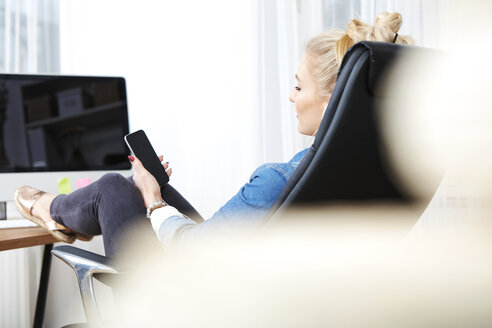 Blond woman sitting at desk with her smartphone listening music - SEGF000519