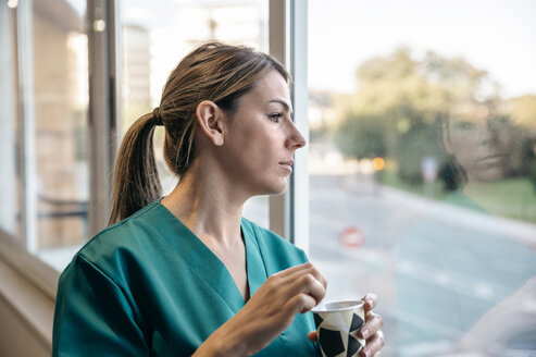 Woman in scrubs having coffee break looking out of the window - DAPF000068