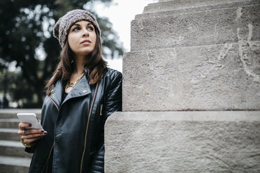 Young woman holding cell phone looking around outdoors - JRFF000522