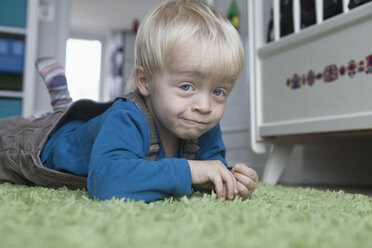 Porträt eines lustigen kleinen blonden Jungen, der auf einem grünen Teppich zu Hause liegt - RBF004244