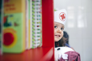 Kleines Mädchen mit Krankenschwesternmütze und Puppe schaut hinter einem Bücherregal hervor - DAPF000086