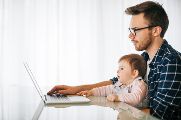Young man with baby girl on his lap trying to work - BRF001298