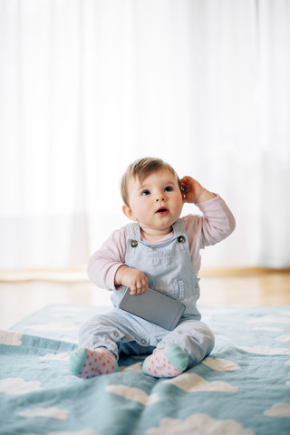 Porträt eines kleinen Mädchens, das auf einer Decke sitzt und ein Smartphone hält, lizenzfreies Stockfoto