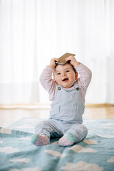 Portrait of baby girl sitting on blanket holding smartphone on the head - BRF001304