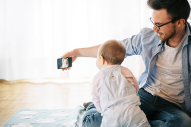 Father sitting on blanket with his little daughter taking selfie - BRF001306