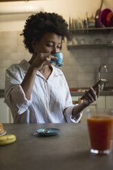 Portrait of young woman drinking espresso in her kitchen while looking at phablet - MAUF000423