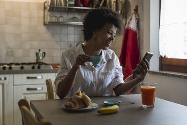 Lachende junge Frau mit Espressotasse, die in ihrer Küche sitzt und auf ihr Phablet schaut - MAUF000425