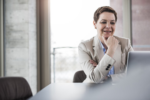 Lächelnde Geschäftsfrau am Schreibtisch mit Blick auf den Laptop, lizenzfreies Stockfoto