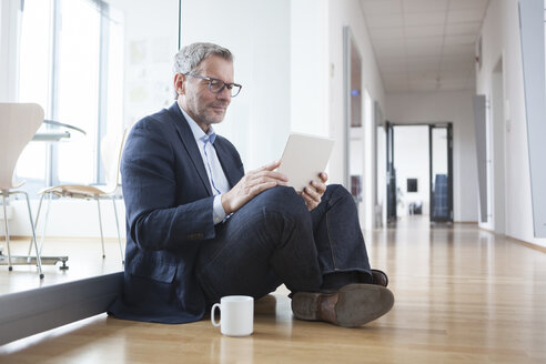 Erfolgreicher Geschäftsmann sitzt auf dem Boden und benutzt ein digitales Tablet in seinem Büro - RBF004327