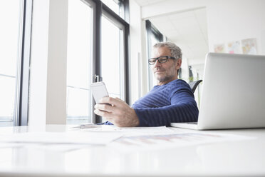 Mature man sitting in office with laptop, using smart phone - RBF004341