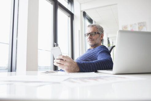 Älterer Mann sitzt im Büro mit Laptop und benutzt ein Smartphone - RBF004341