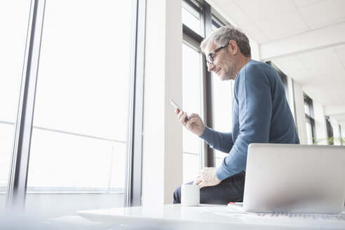 Mature man sitting in office with laptop, using smart phone - RBF004345