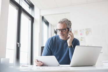 Älterer Mann sitzt im Büro und benutzt Tablet-PC und Smartphone - RBF004347