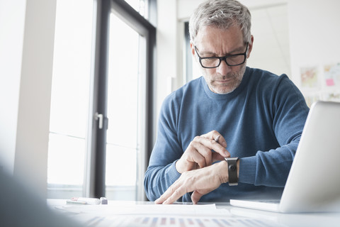 Älterer Mann sitzt im Büro und arbeitet mit Laptop und Smartwatch, lizenzfreies Stockfoto