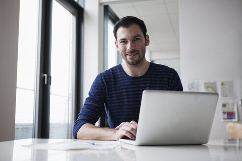 Junger Mann sitzt im Büro und benutzt einen Laptop - RBF004406