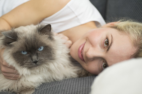 Porträt einer lächelnden jungen Frau, die mit ihrer Katze auf der Couch liegt, lizenzfreies Stockfoto