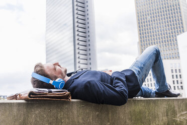 Young businessman lying on wall listening music with headphones - UUF006885