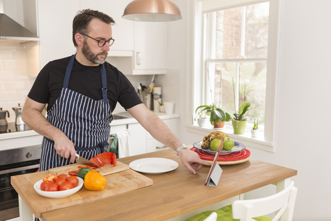 Mann benutzt digitales Tablet in seiner Küche bei der Zubereitung von Gemüse, lizenzfreies Stockfoto