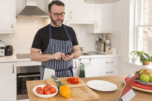 Porträt eines Mannes, der ein Smartphone benutzt, während er in der Küche Essen zubereitet - BOYF000253