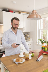 Mann steht morgens in seiner Küche und schaut auf sein Smartphone - BOYF000265