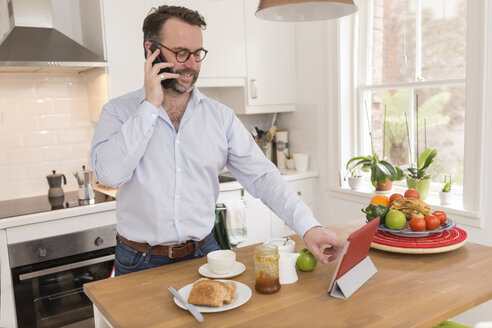 Mann steht am Frühstückstisch in der Küche und telefoniert mit einem Smartphone, während er ein digitales Tablet benutzt - BOYF000269