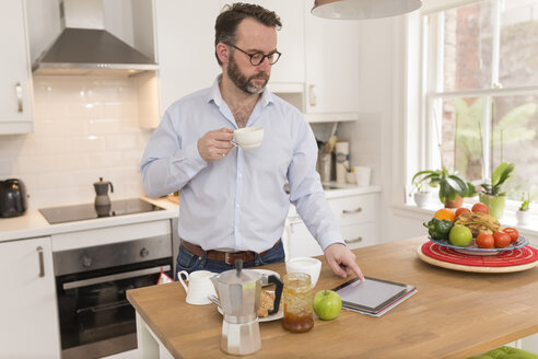 Mann mit Tasse Kaffee in der Küche stehend und mit digitalem Tablet - BOYF000278