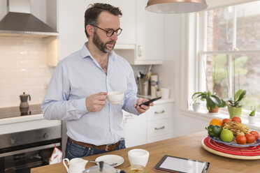 Mann steht in der Küche mit einer Tasse Kaffee und schaut auf sein Smartphone - BOYF000279