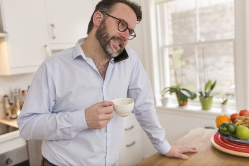 Mann steht in der Küche mit Tasse und telefoniert mit Smartphone - BOYF000280