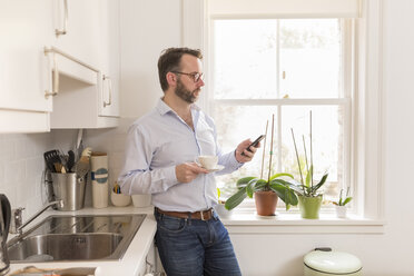 Mann steht in der Küche mit einer Tasse Kaffee und schaut auf sein Smartphone - BOYF000281