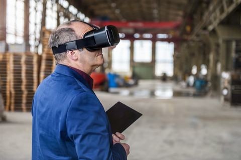 Manager steht in einer Fabrikhalle und schaut durch eine VR-Brille, lizenzfreies Stockfoto