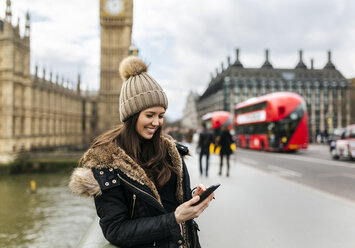 Großbritannien, London, lächelnde junge Frau schaut auf ihr Smartphone vor dem Palace of Westminster - MGOF001730